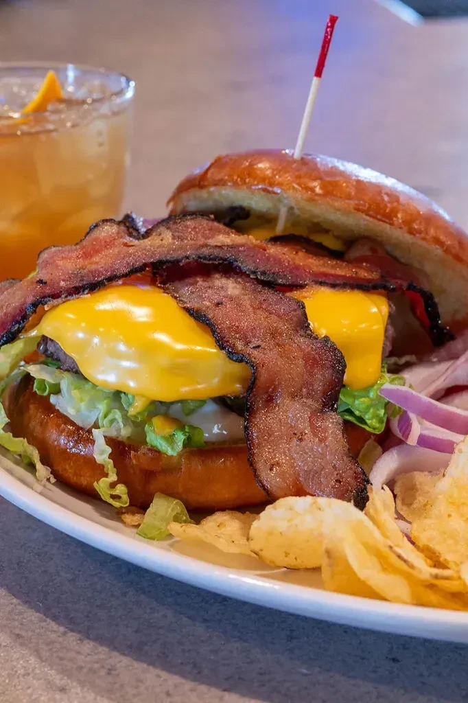 Close-up burger with melted cheese, bacon, lettuce, chips, and a drink.