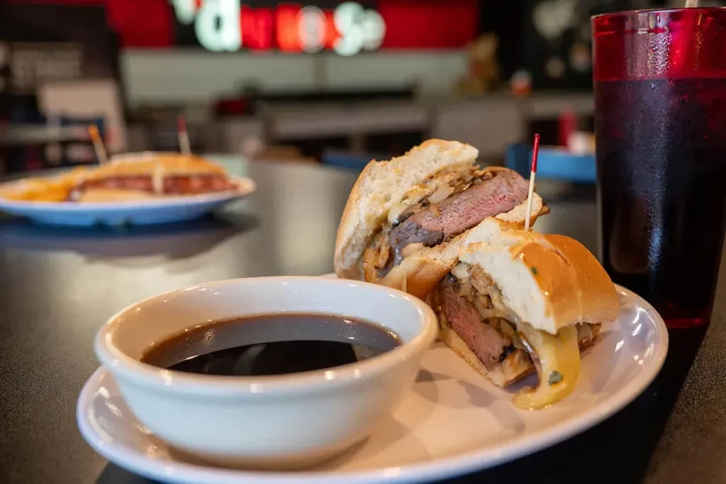 Medium-rare roast beef sandwich with melted cheese, au jus, and iced drink.