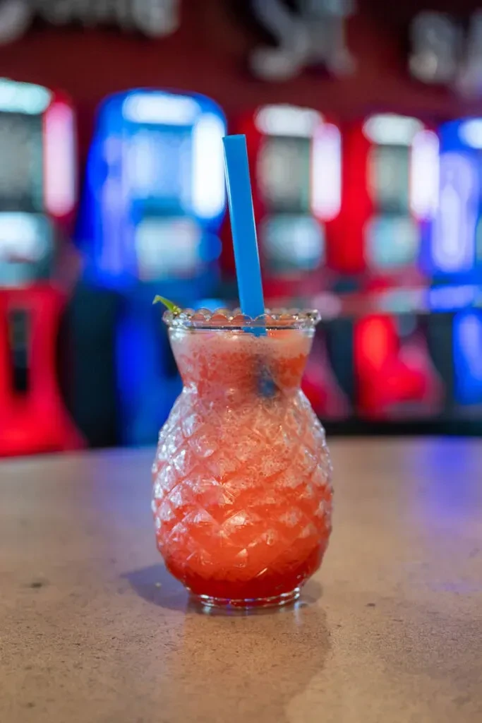Red-pink frosty drink in pineapple glass, blue straw, green garnish.