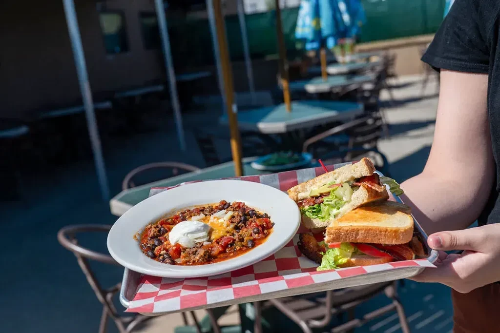 Person holding metal tray with chili and BLT sandwich outdoors.