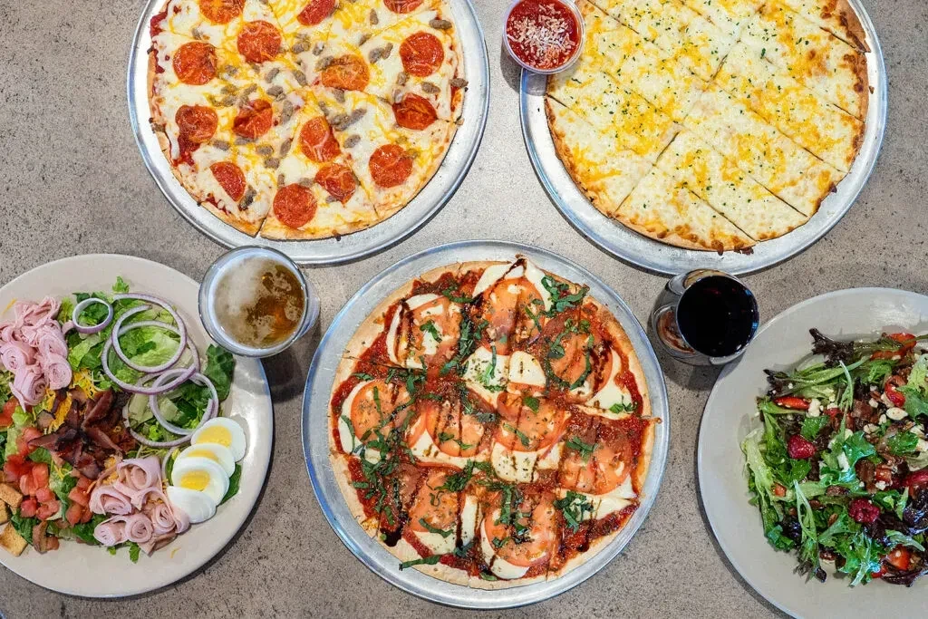 Top-down view of three pizzas, two salads, beer, and wine.
