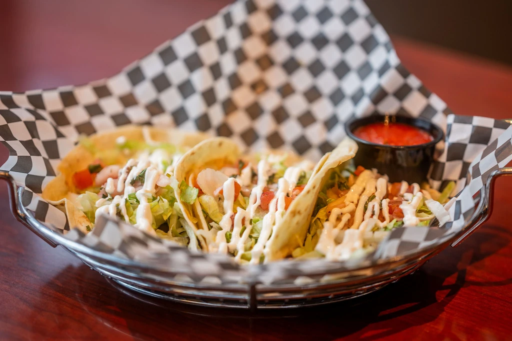 Three shrimp tacos, lettuce, tomato, creamy sauce, and salsa in basket.