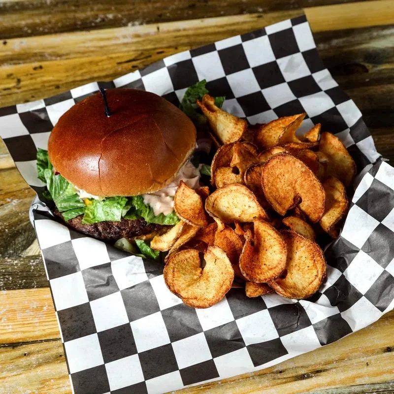 Overhead: Thick burger, crispy chips on checkered paper and rustic wood.