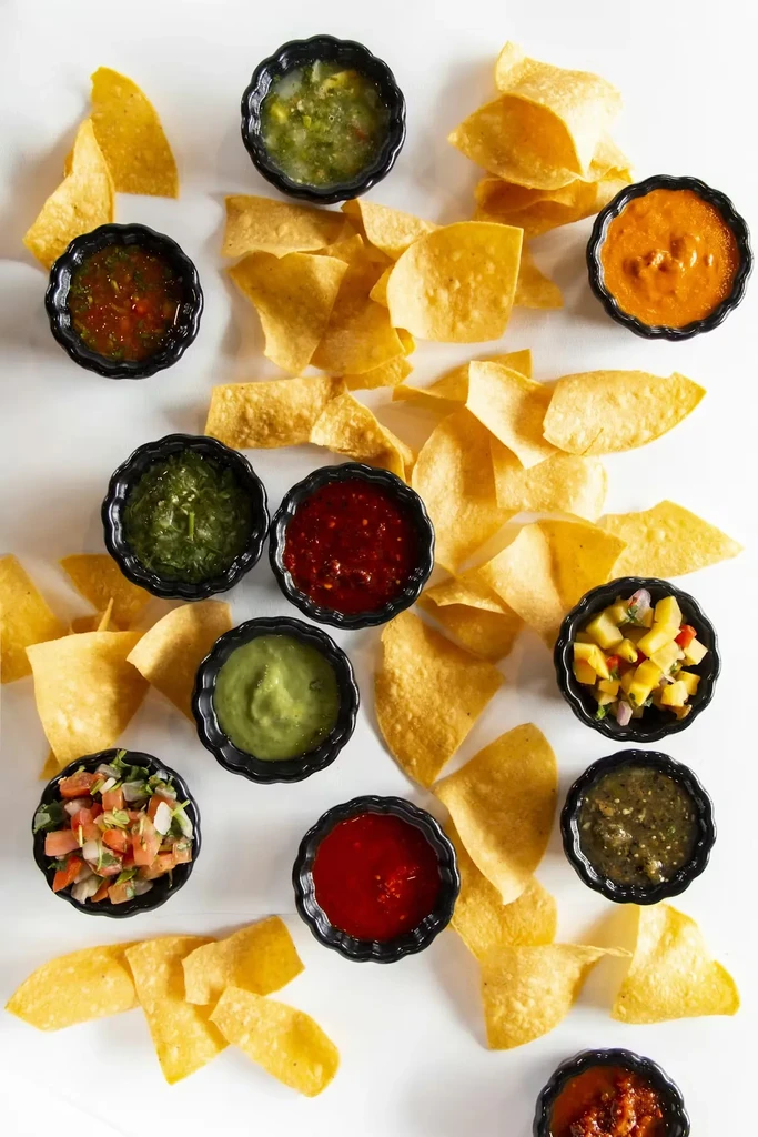 Golden tortilla chips surround ten bowls of colorful dips on white.