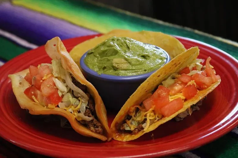 Two hard-shell tacos, guacamole on a red plate, serape background.