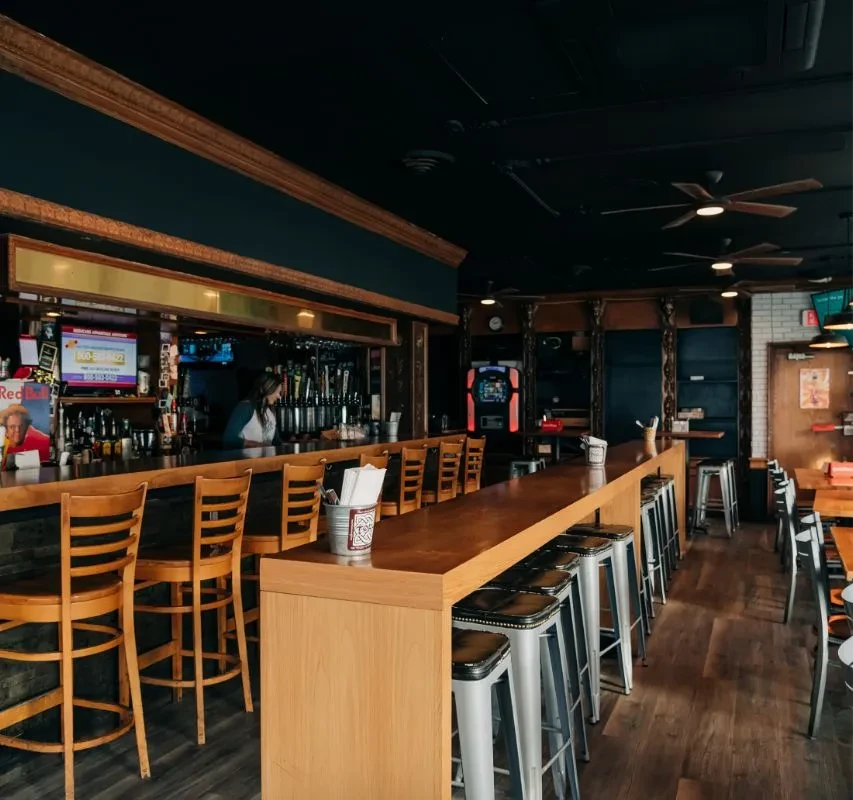 Well-lit modern bar with wooden counter, stools, bottles, taps, and jukebox.