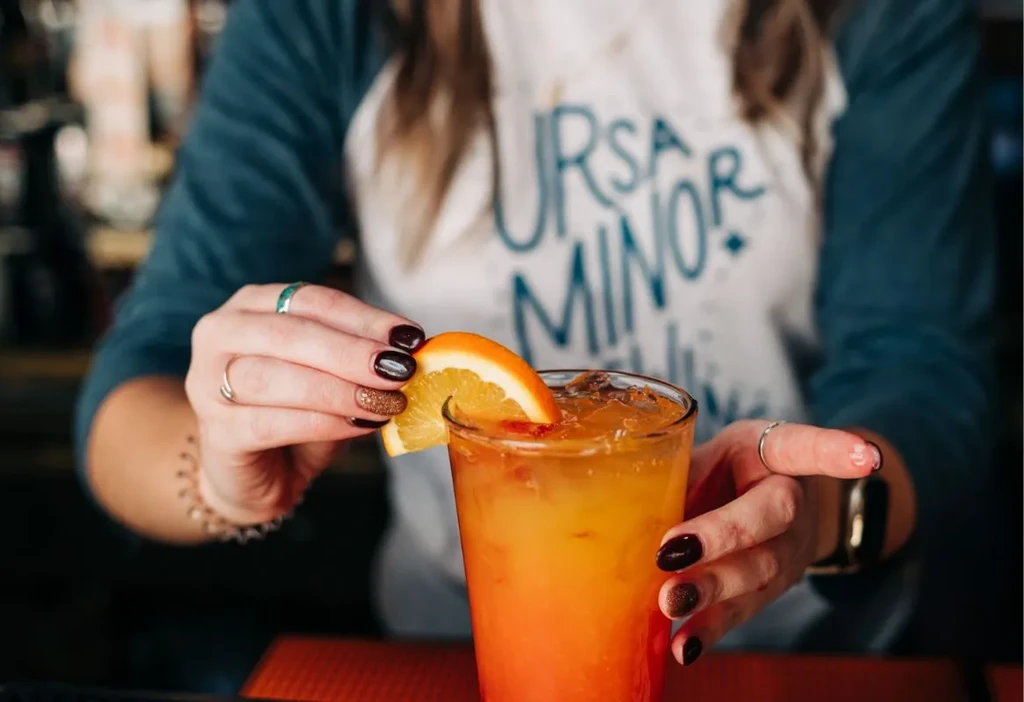 Hands garnish a vibrant orange and yellow cocktail with an orange slice.