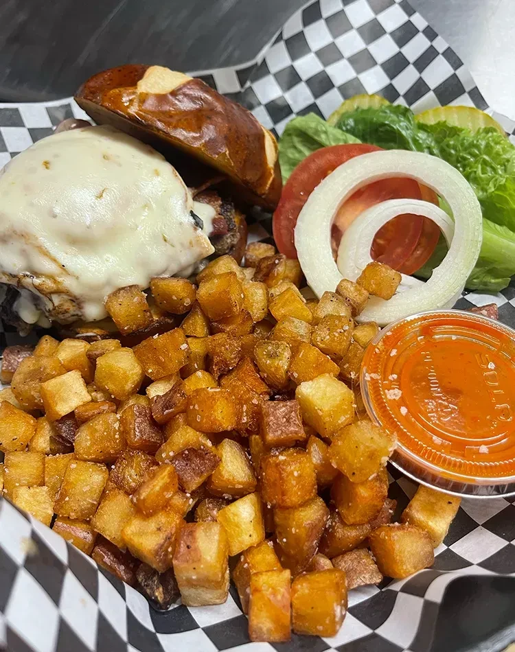 Cheesy pretzel bun burger, diced potatoes, vegetables, and sauce in basket.