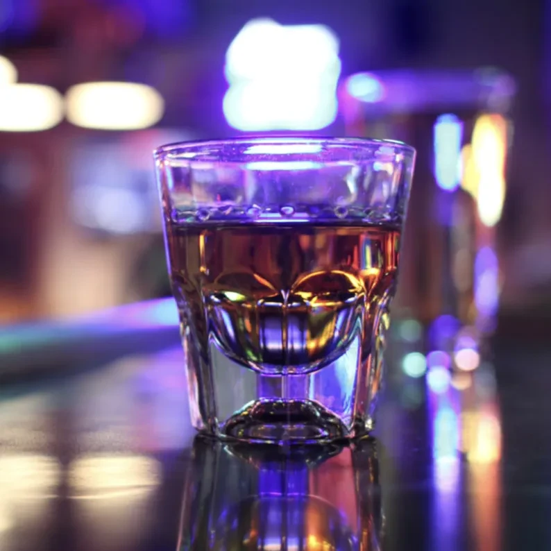 Faceted shot glass with amber liquid, reflecting colorful bokeh lights.
