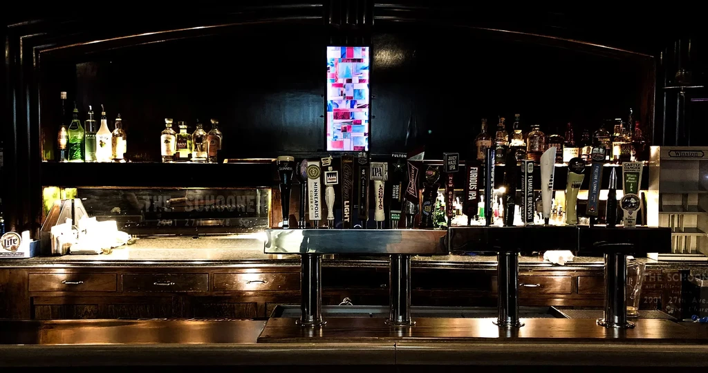 Dimly lit bar counter with beer taps, liquor bottles, and abstract digital screen.