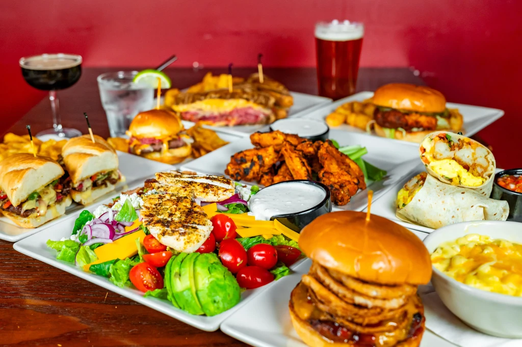 Extensive table spread of burgers, wings, subs, mac and cheese, drinks.