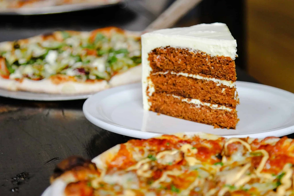 Close-up of carrot cake slice and two pizzas on a dark table.