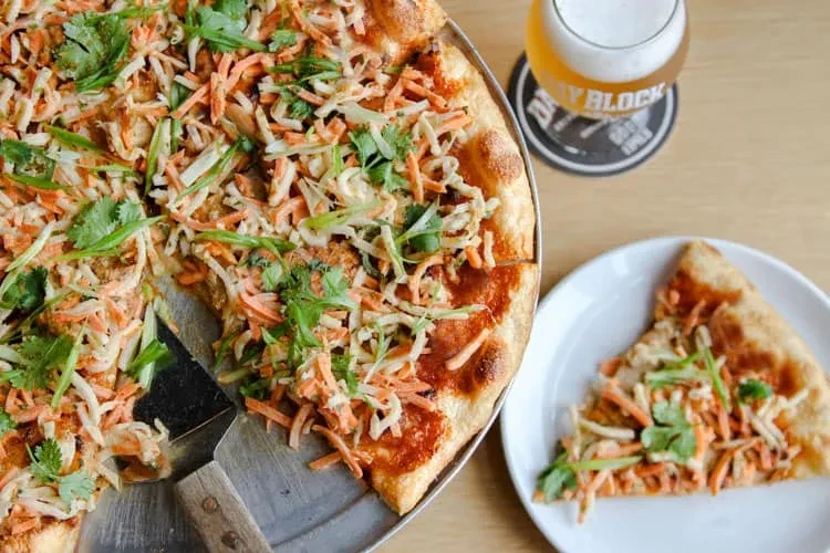 Pizza with shredded carrots, cabbage, cilantro; a removed slice and beer.