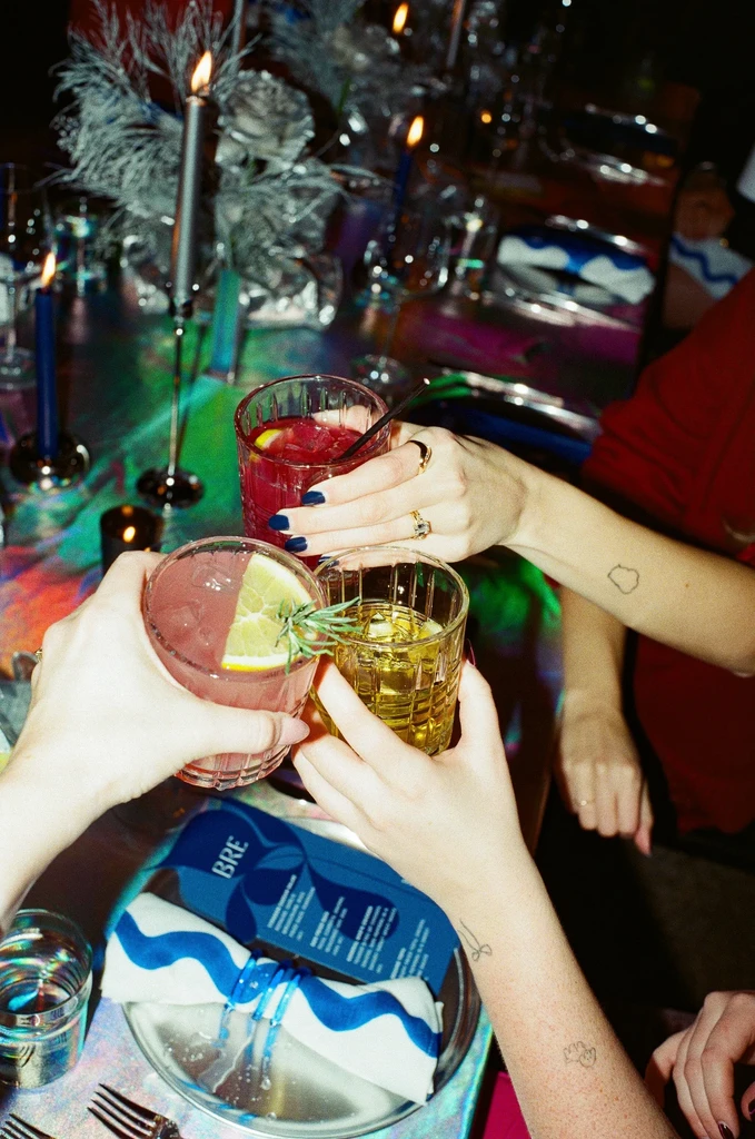 Four hands clinking three colorful cocktails on an iridescent table.