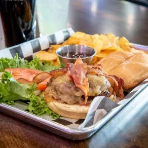 Burger with cheese, bacon, lettuce, tomato, chips, and dipping sauce.