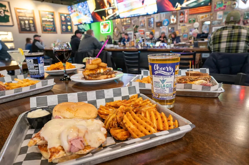 Lively pub table with a sandwich, fries, beer, and blurred patrons.