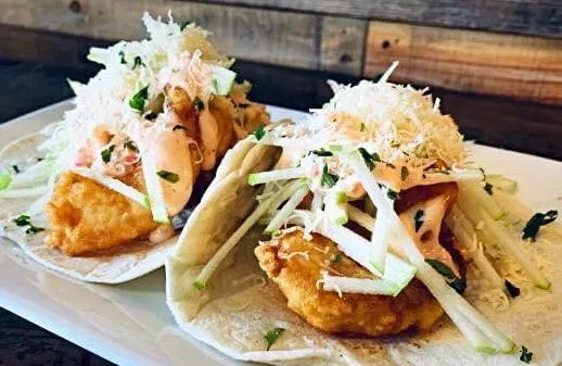 Two crispy fried fish tacos with slaw, sauce, and herbs on a plate.