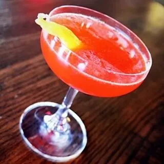 Vibrant red cocktail in a coupe glass with yellow peel on wood.
