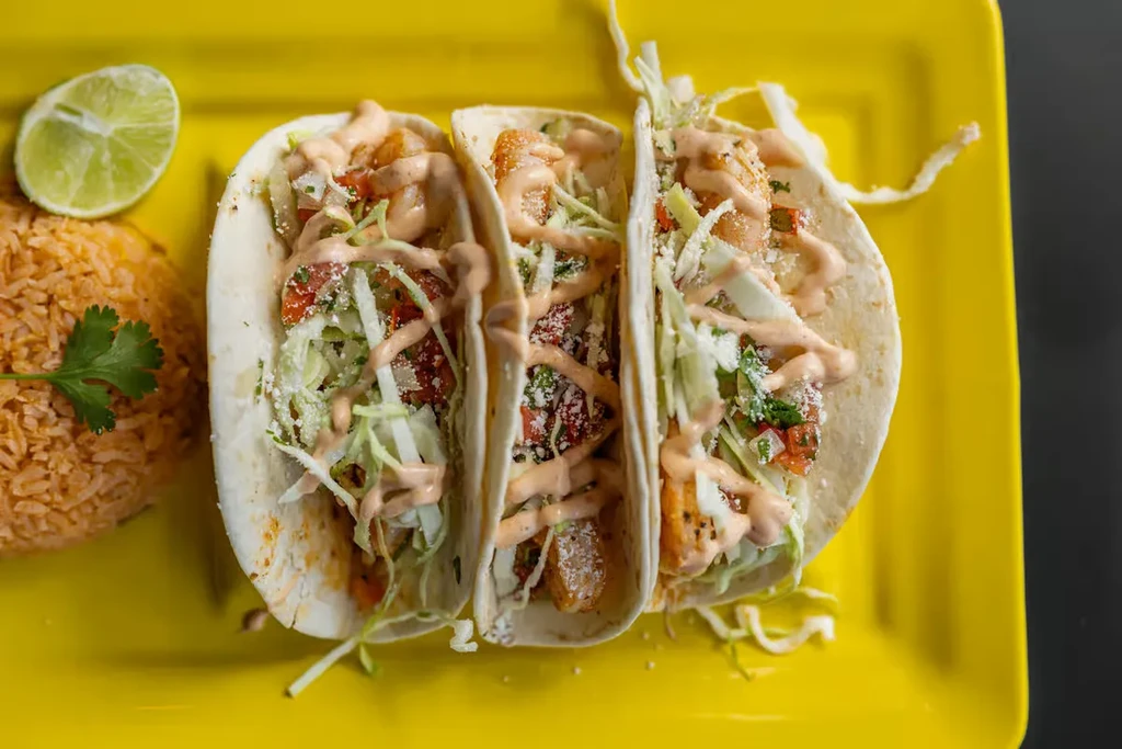 Three soft-shell tacos with rice and lime on a yellow plate.