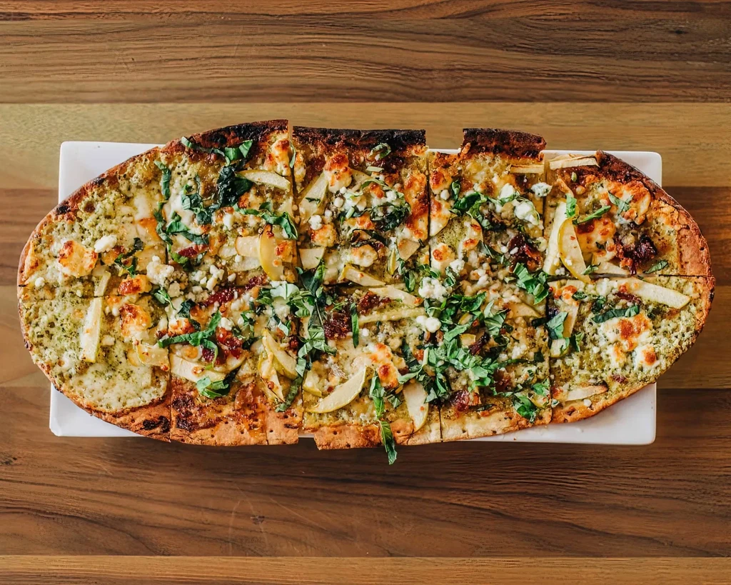 Top-down pesto flatbread with fruit, cheese, and herbs on a wooden table.