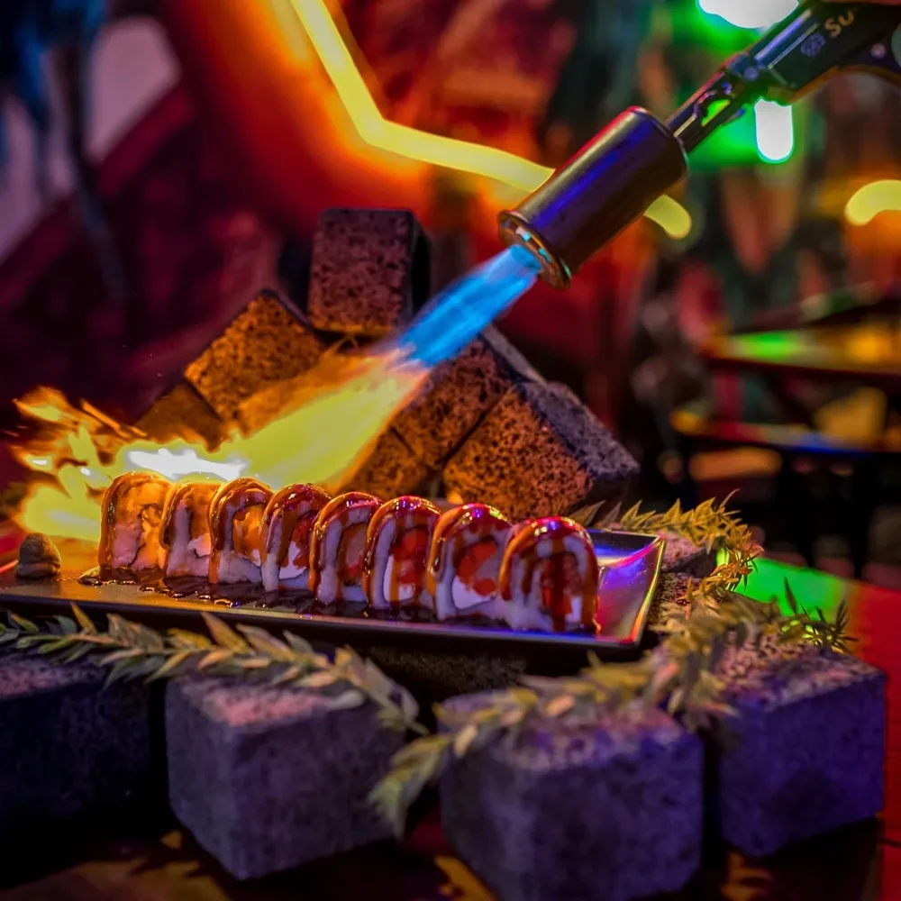 Sushi rolls on dark dish, actively torched by blue flame.