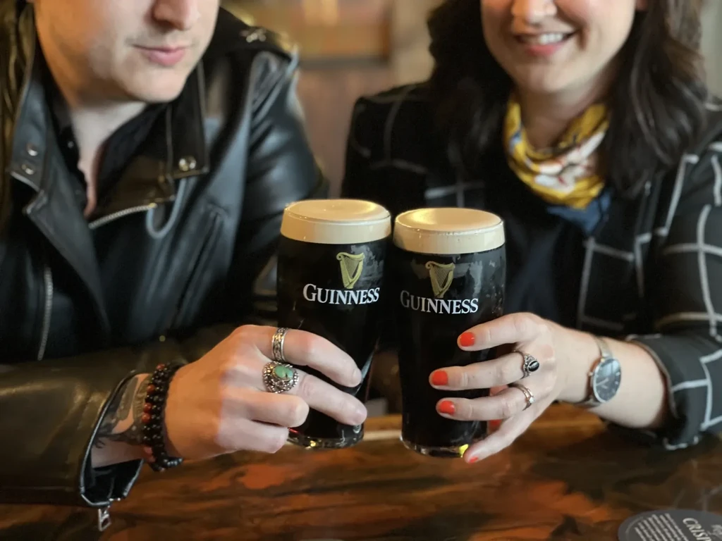 Two people clink Guinness pint glasses in a warm pub.