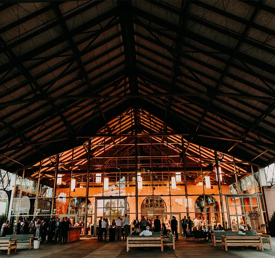 Warm industrial venue with truss roof, glass facade, and socializing people.