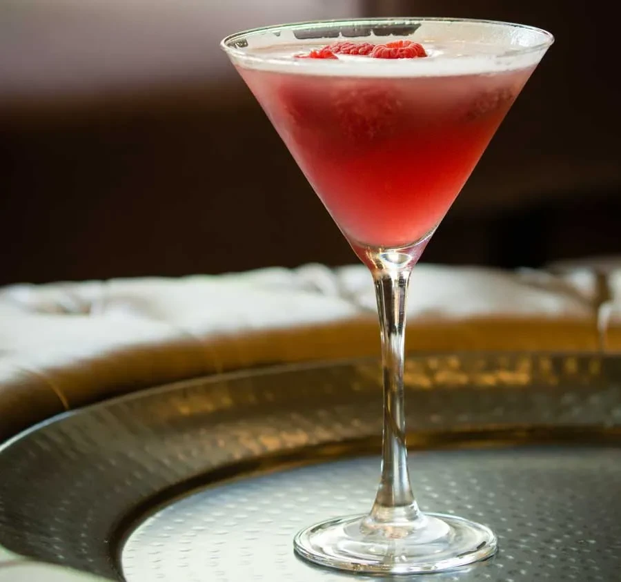 Reddish-pink cocktail in martini glass with raspberries and frothy top.
