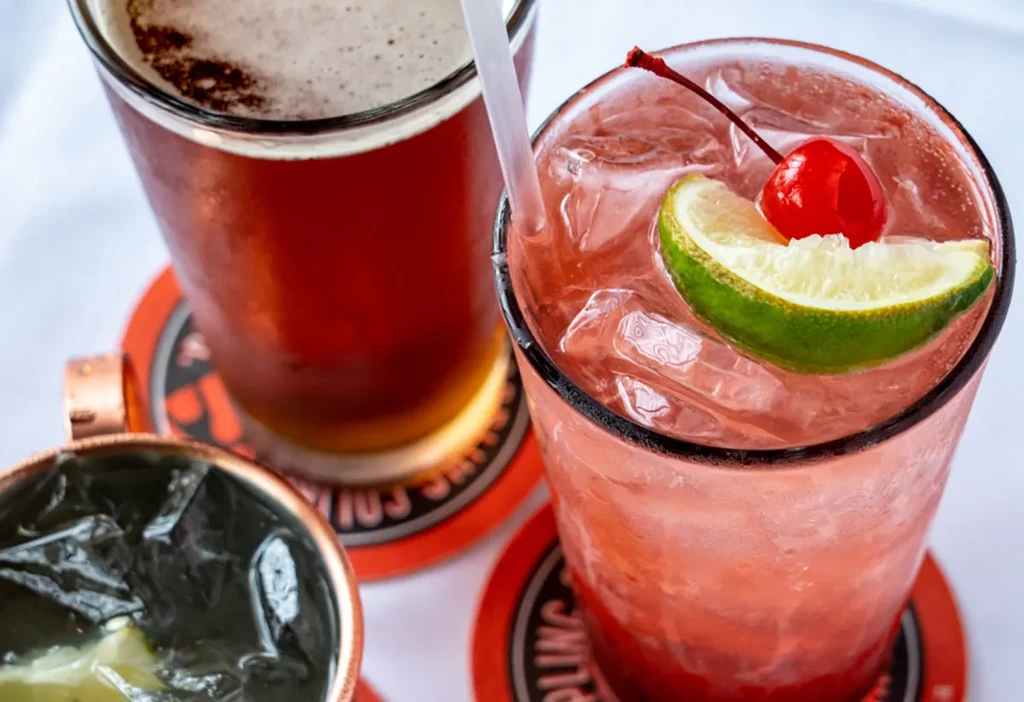 Pink cocktail, amber beer, and copper mug on orange coasters.