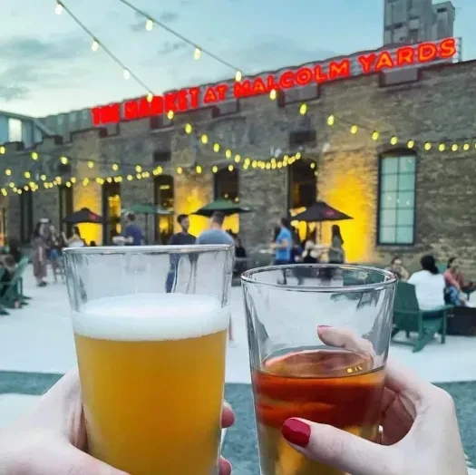 Two hands toasting beer and a drink at Malcolm Yards.