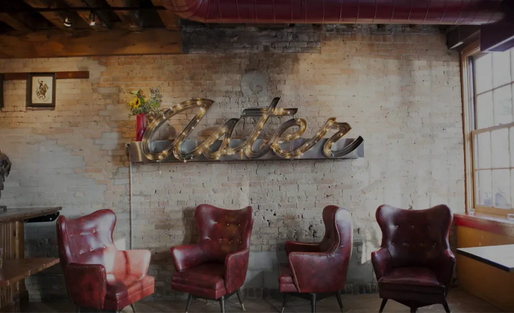 Rustic-industrial restaurant with lighted "Cater" sign and red leather chairs.