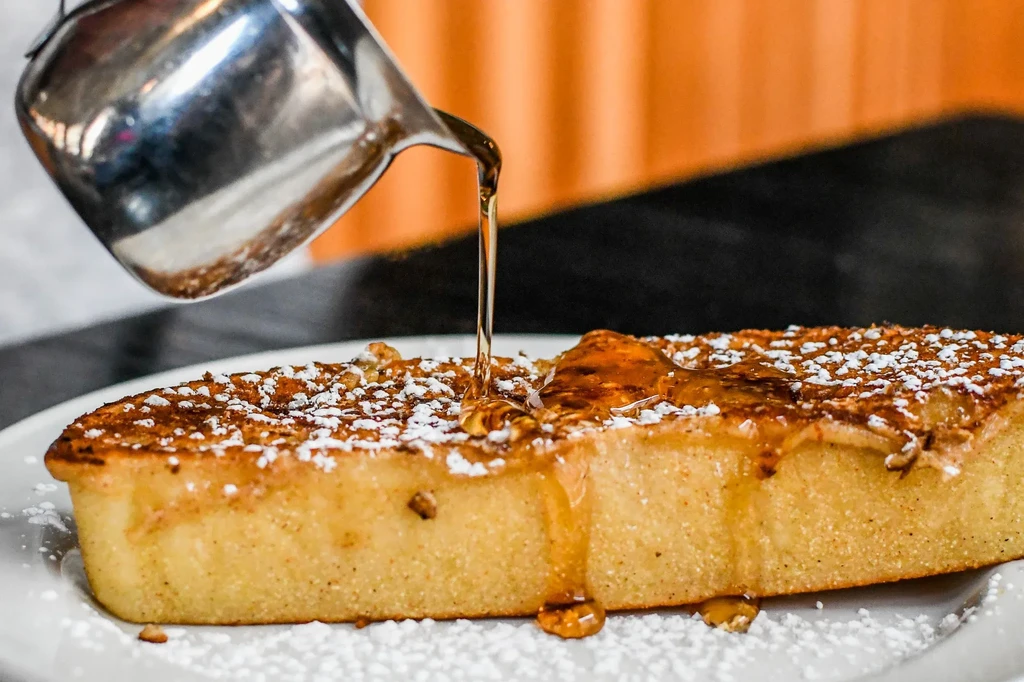 Thick French toast with powdered sugar, nuts, and golden syrup pouring.