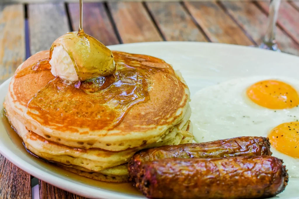 Pancakes with pouring syrup, fried eggs, and sausage on a plate.