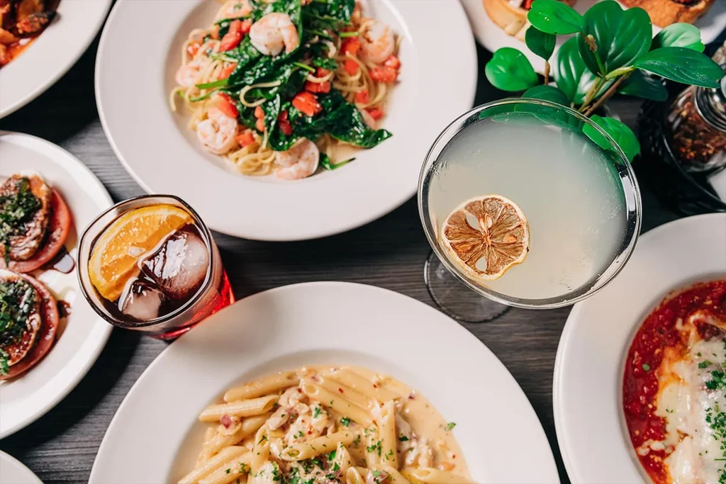 Plates of pasta, appetizers, baked dish, and cocktails on a dark table.