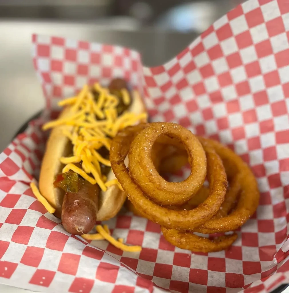 Hot dog with cheese, relish, and curly fries in a checkered basket.