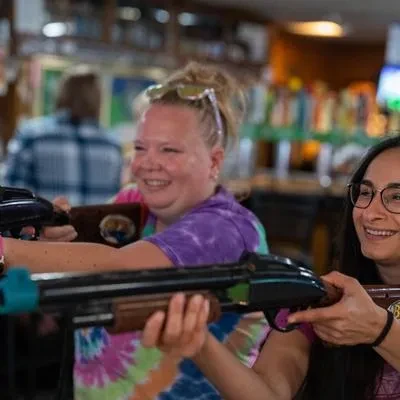 Two smiling women hold toy shotguns in a lively bar.