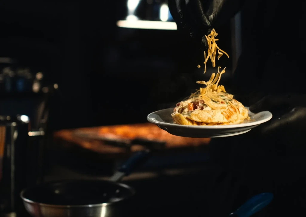 Close-up: Gloved hand sprinkles cheese onto a dish, kitchen background.