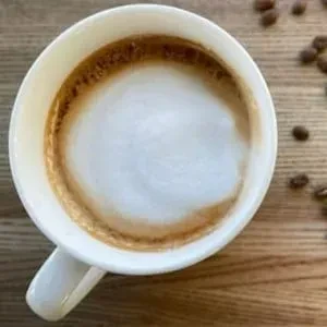 White mug of foamy coffee on wood, with scattered coffee beans.