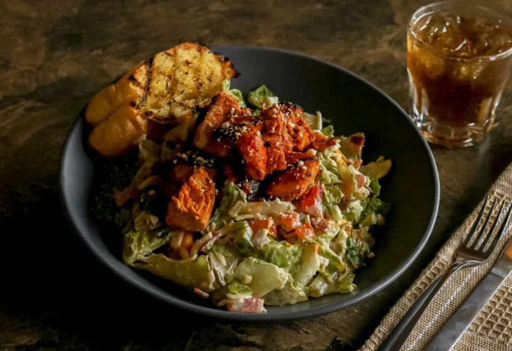 Salad bowl with sauced chicken, grilled bread, and an amber drink.