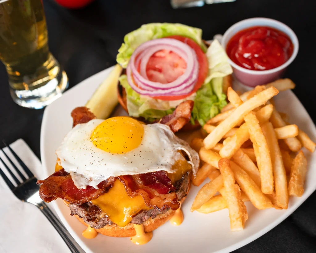 Burger with melted cheese, bacon, runny egg, fries, and ketchup.