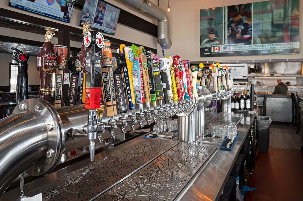 Numerous beer taps on a stainless steel bar with sports TVs.