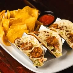 Three shrimp tacos, tortilla chips, and red salsa on a plate.