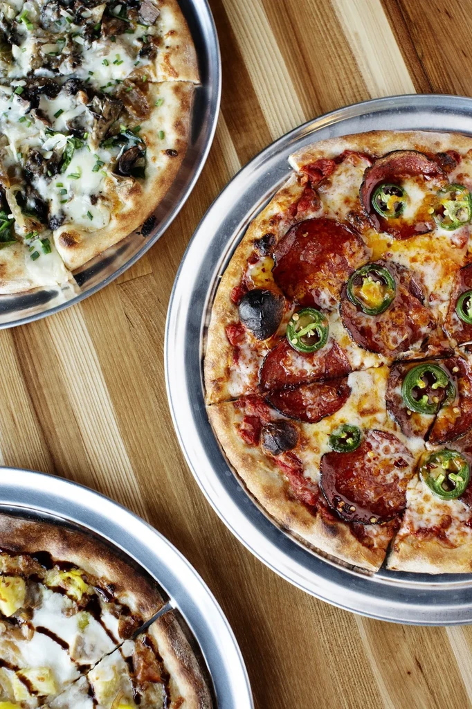 Three different pizzas on silver trays on a wooden table.