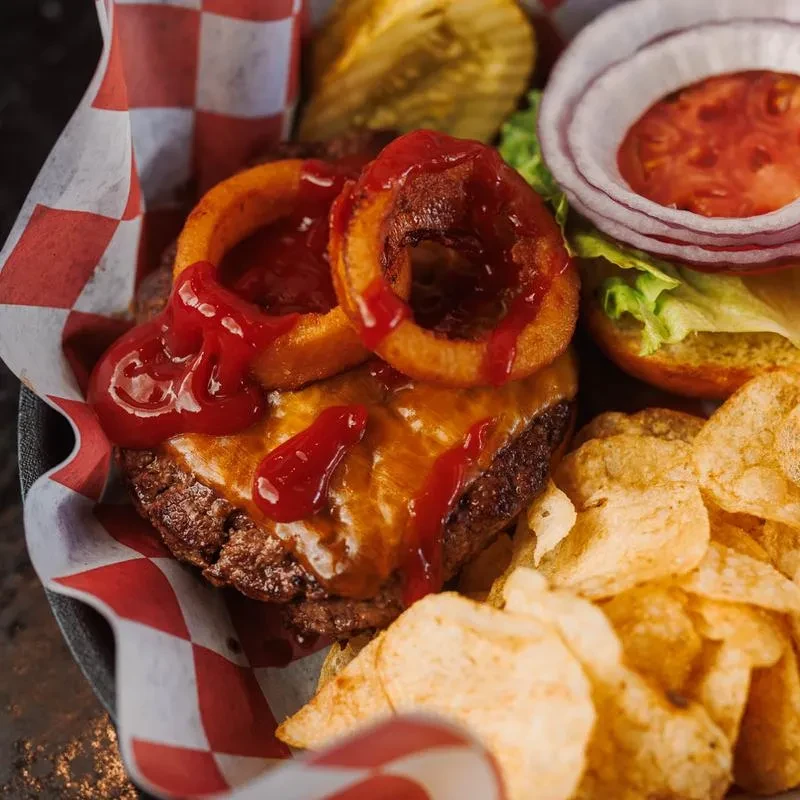 Gourmet burger with onion rings, cheese, chips, pickle, in a checkered basket.