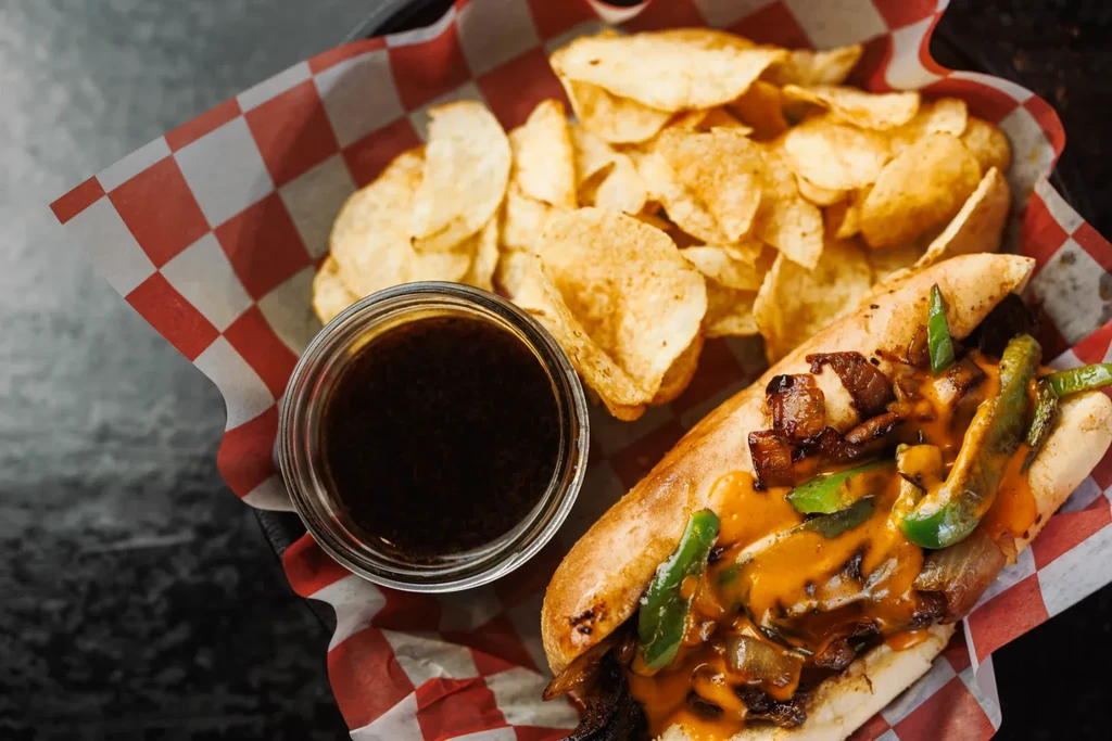 A cheesesteak sandwich, potato chips, and au jus in a checkered basket.