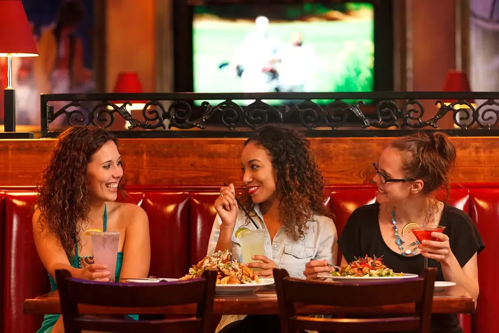 Three smiling women enjoy drinks and food in a restaurant.