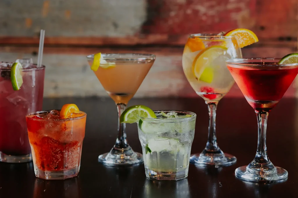 Six vibrant cocktails with ice and fruit on a dark reflective surface.