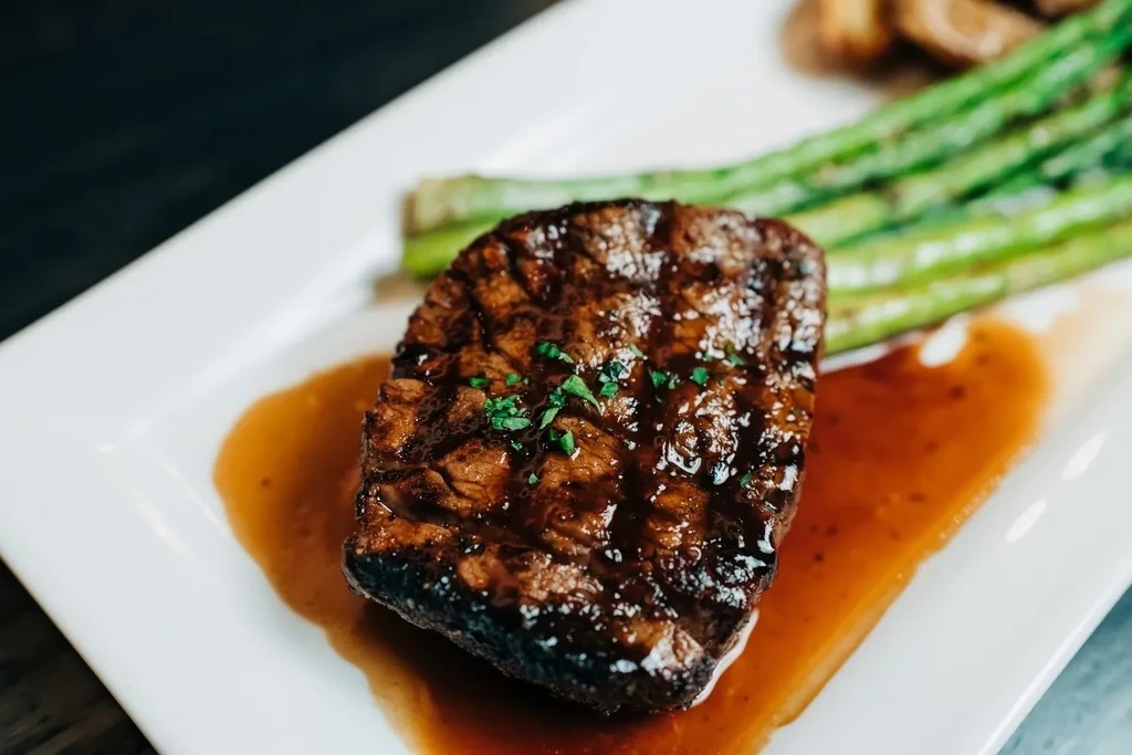 Seared meat, green herbs, amber sauce, asparagus, on a white plate.