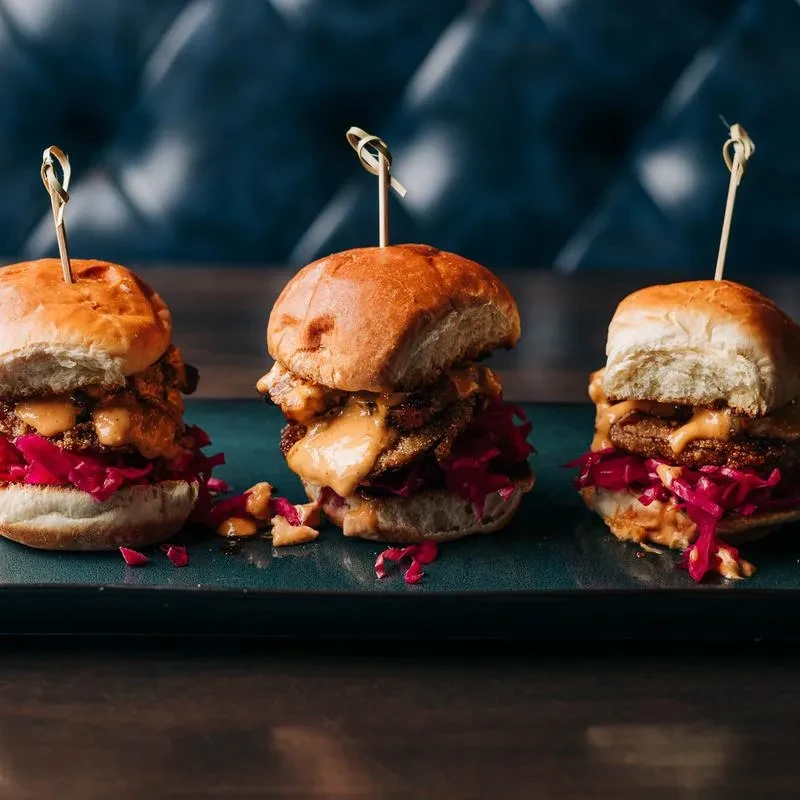 Three sliders with red cabbage slaw and creamy sauce on a dark platter.