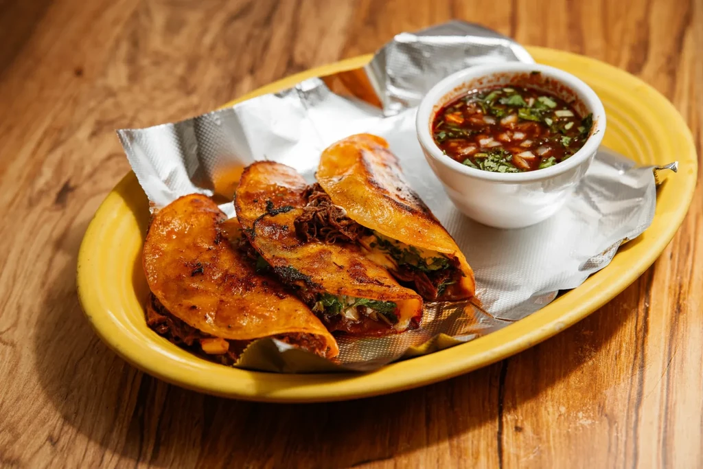 Three crispy birria tacos with meat, cilantro, and a side of consommé.